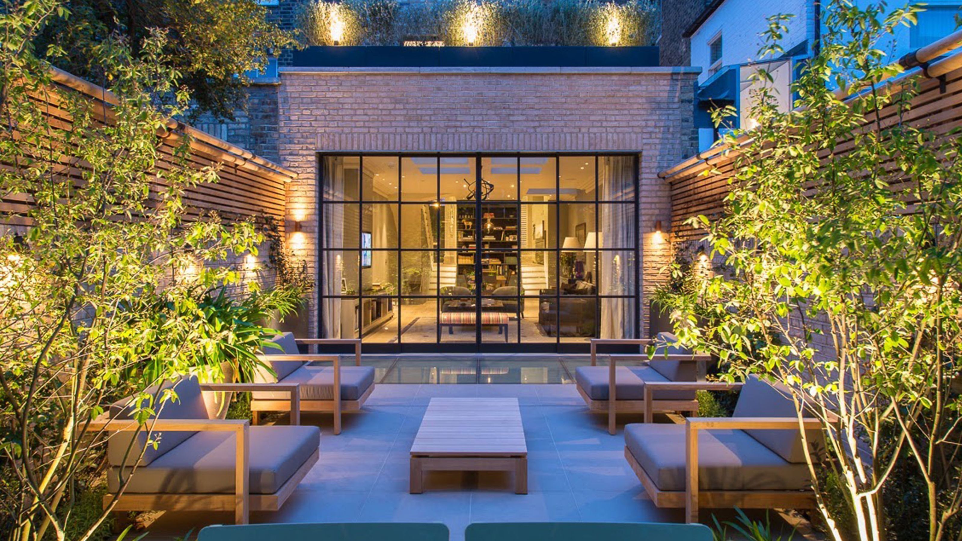 An urban modern garden, with a view of the home through the crittal style doors, with two large arm chairs either side of a garden coffee table with lit trees and fence panels all around