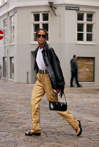 a woman wearing a leather fringe jacket with a white t-shirt, studded belt, silk pants and sandals