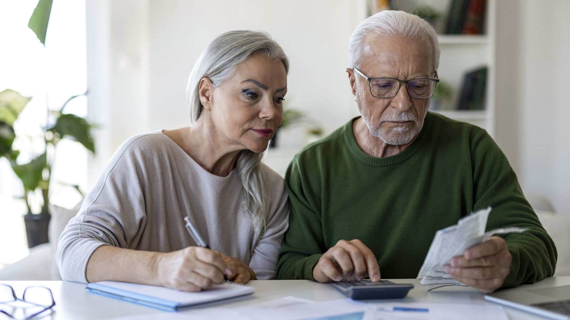 A serious-looking older couple work on financial planning at their kitchen counter.