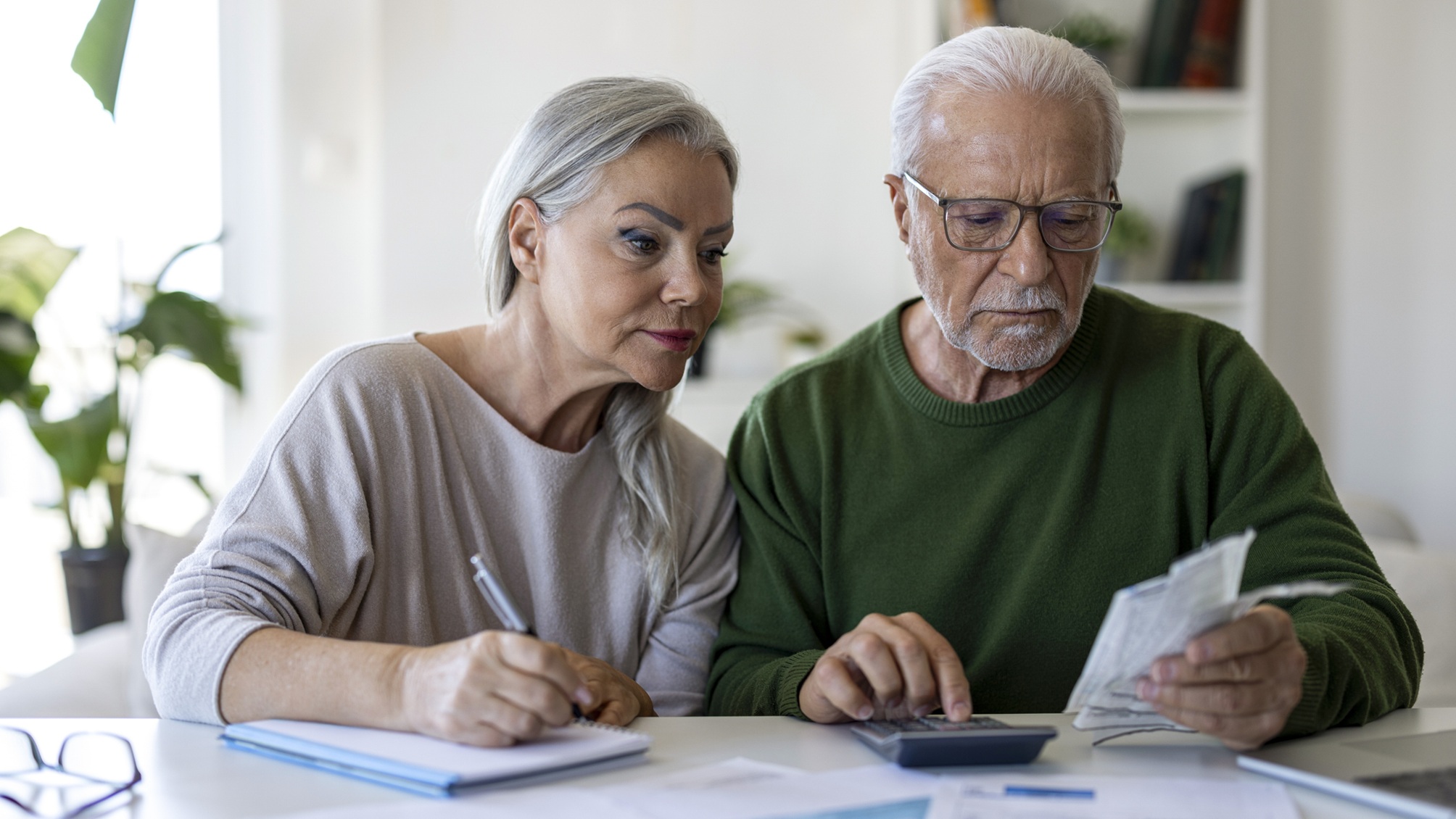 A serious-looking older couple work on financial planning at their kitchen counter. 
