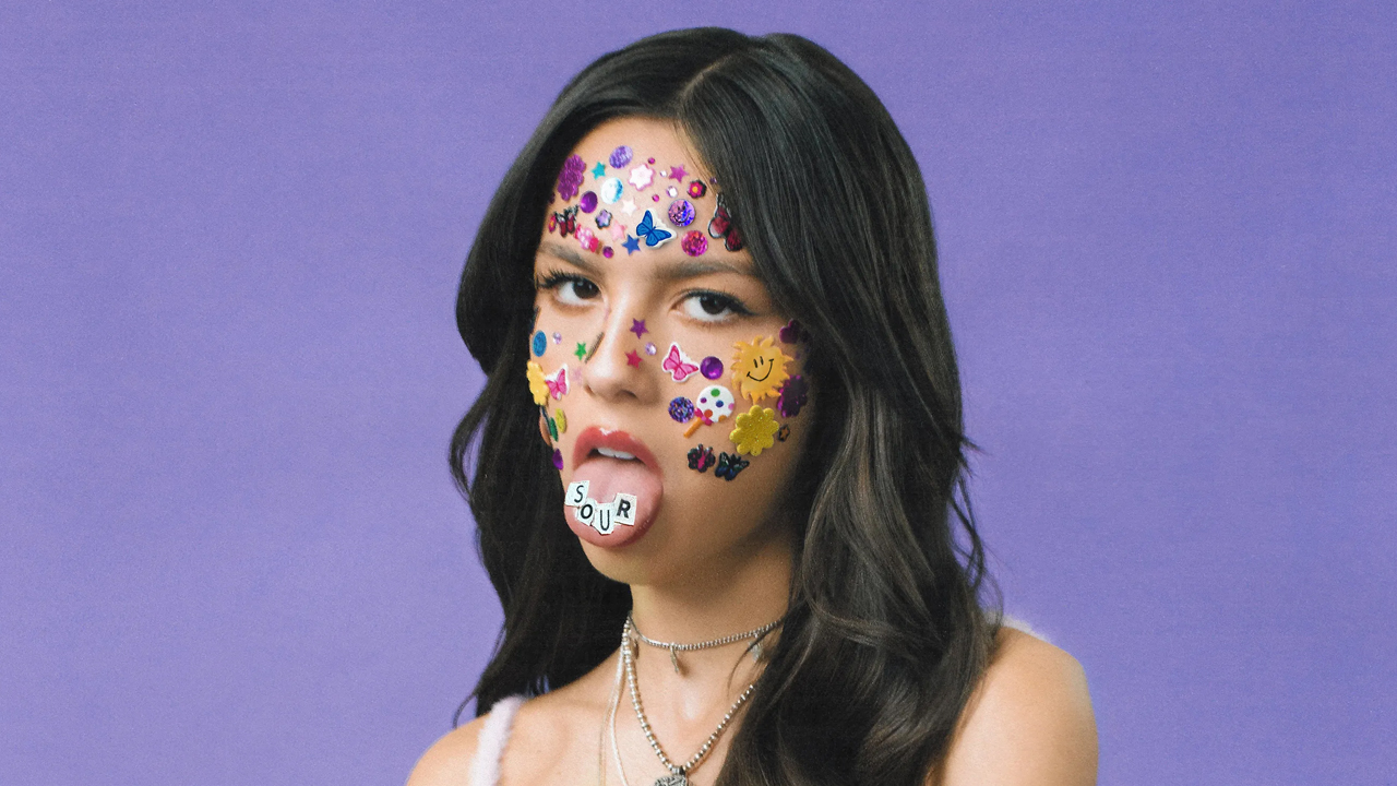 olivia rodrigo sticking her tongue out, with stickers on her face and tongue that spell the word 'sour'