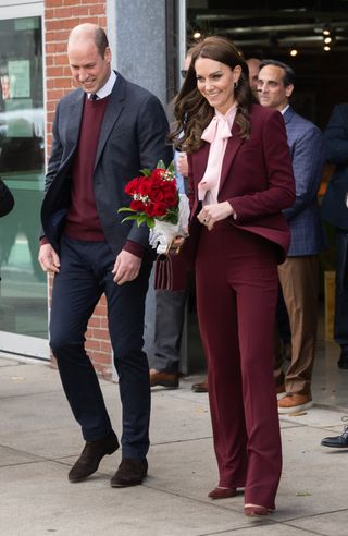 Kate Middleton wearing a maroon suit and pink blouse holding flowers walking next to Prince William leaving a building