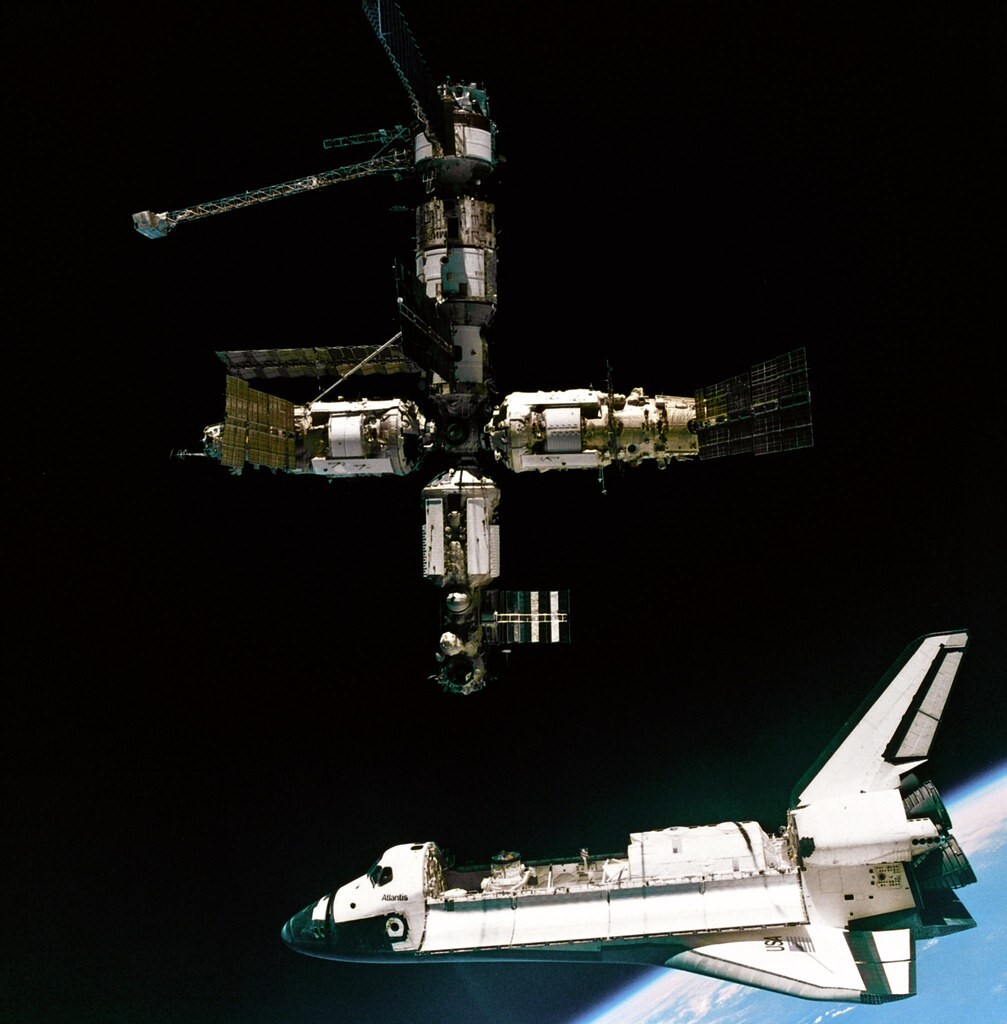 A space shuttle with cargo bay open is docked with a cross-sectional space station.