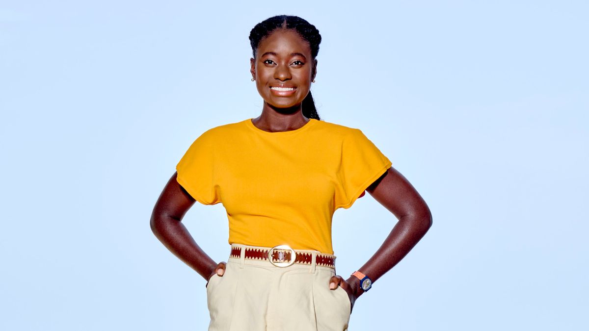 A generic shot of Shantol Jackson as DS Naomi Thomas in Death in Paradise. She is standing in front of a clear blue sky, wearing a mustard-yellow top with high-waisted cream-coloured trousers. She is smiling brightly and has her hands in her pockets.