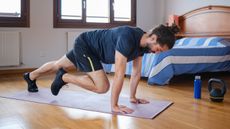 A man performs mountain climbers in his bedroom on an exercise mat. He is on hands hands, arms straight, with one leg straight behind him and one leg bent close to his chest. 
