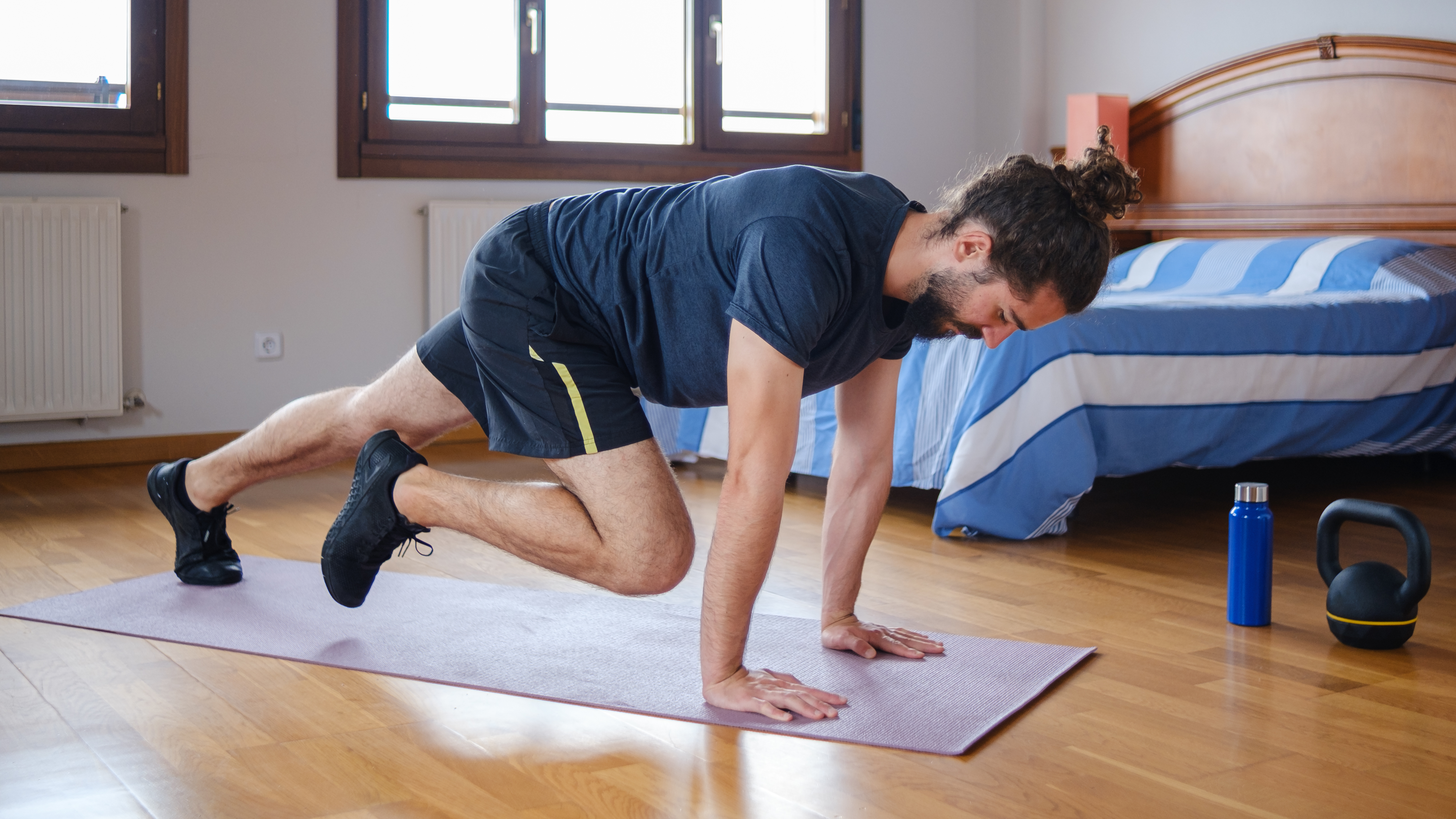 A man performs mountain climbers in his bedroom on an exercise mat. He is on hands hands, arms straight, with one leg straight behind him and one leg bent close to his chest. 