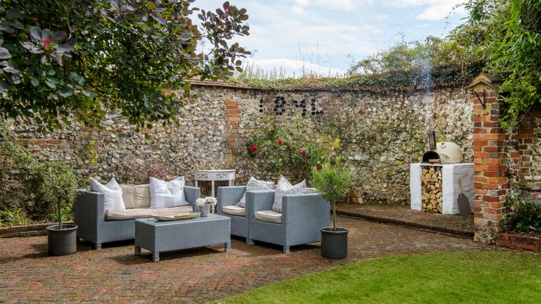 Garden wall ideas featuring a character flint wall with an outdoor pizza oven, climbing plants and a gray outdoor sofa set.