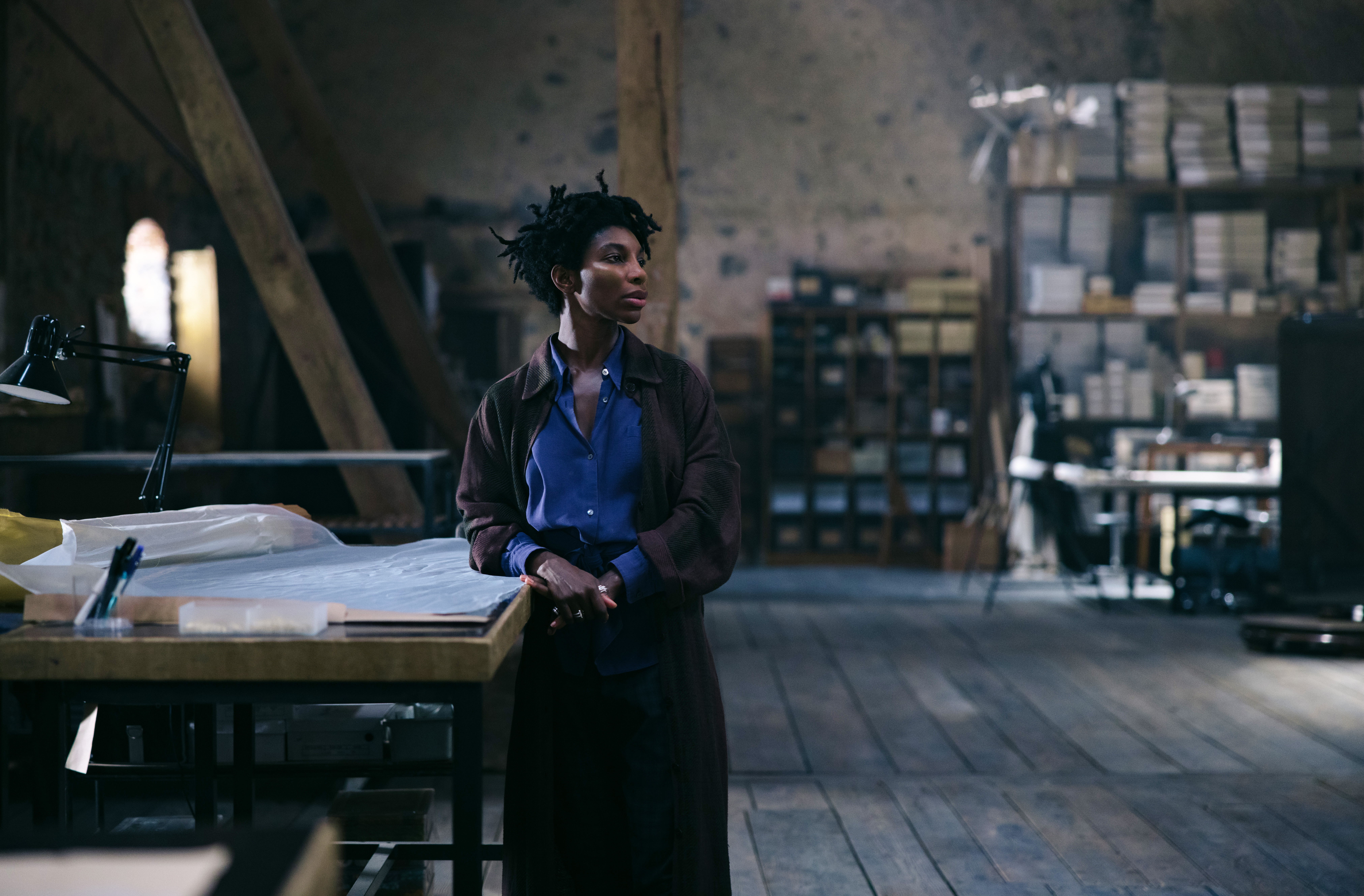 A still from the movie 'Mother Mary' of Michaela Cole wearing a blue shirt, brown jacket, black pants on set.