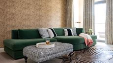 A living area with brown burl wallpapered walls, a deep green curved velvet couch with black and white patterned throw pillows, a rectangular black and white polka dot ottoman with a circular tray with a ribbed glass