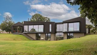 Modern black-clad house with large floor-to-ceiling windows, stone base, and multiple stepped entrances, set on a sloping green lawn under a blue sky.
