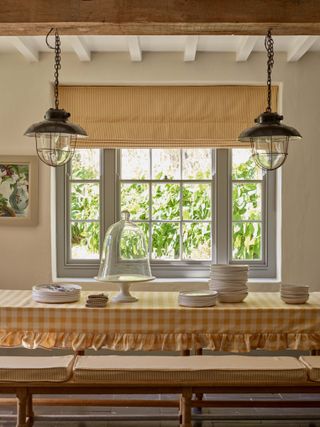 farmhouse spring decor idea with yellow gingham table cloth with frills, yellow stripe bench seats and matching blind, fisherman style pendant lights, beams, artwork, plates and cake stand on table