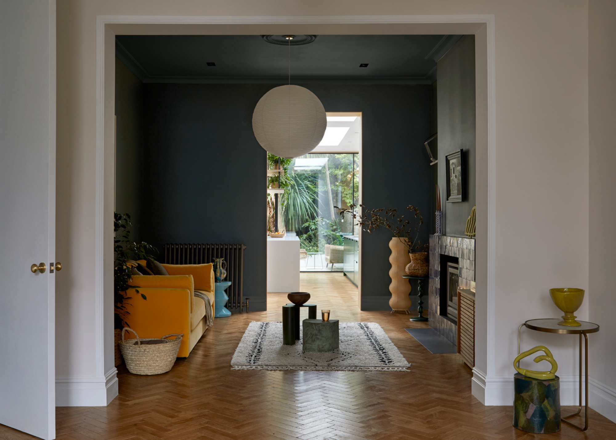 A living room with a pendant rice paper lamp, an orange-yellow sofa, a woven storage basket, a rug, and a hanging painting over a fireplace