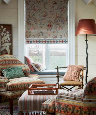 A maximalist living room with a bright patterned arm chair in front of a slim floor-to-ceiling window, covered with a paisley print roman blind.