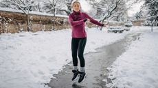 Woman on park path surrounded by snow jumps and swings her arms. She is dressed in black leggings, thick gray socks, black trainers and a reddish-purple long-sleeved top 