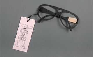 View of the illustration side of ﻿Giles Deacon's pink label invitation which is attached to a pair of black sunglasses made out of paper. The invitation is pictured against a grey background