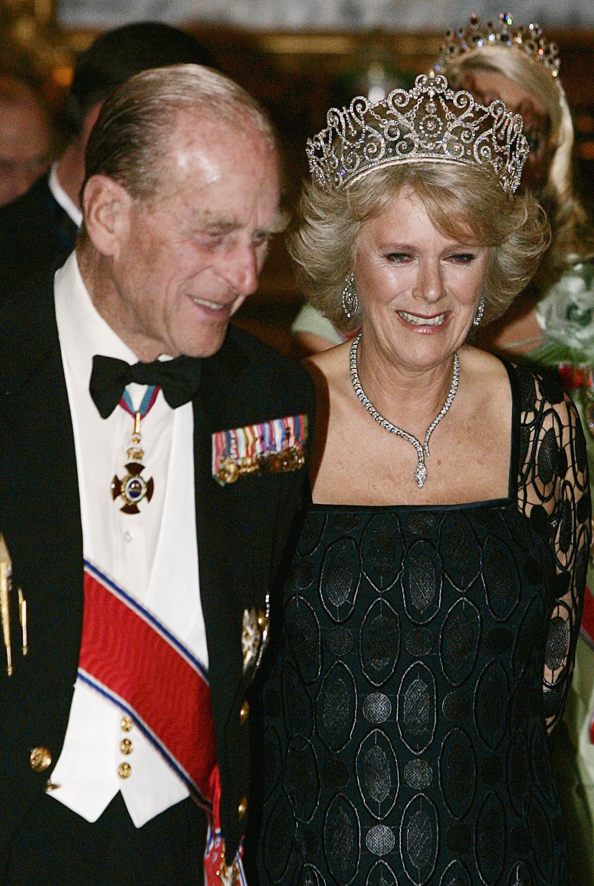 Prince Philip wearing a tuxedo walking next to Queen Camilla wearing a tiara