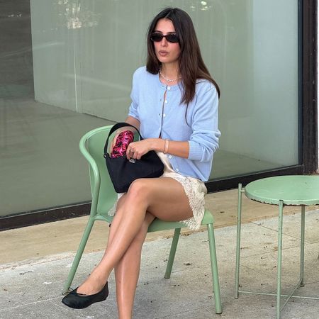 French influencer Tamara Mory wearing a light blue cardigan, cream lace-trimmed miniskirt, and bkack ballet flats