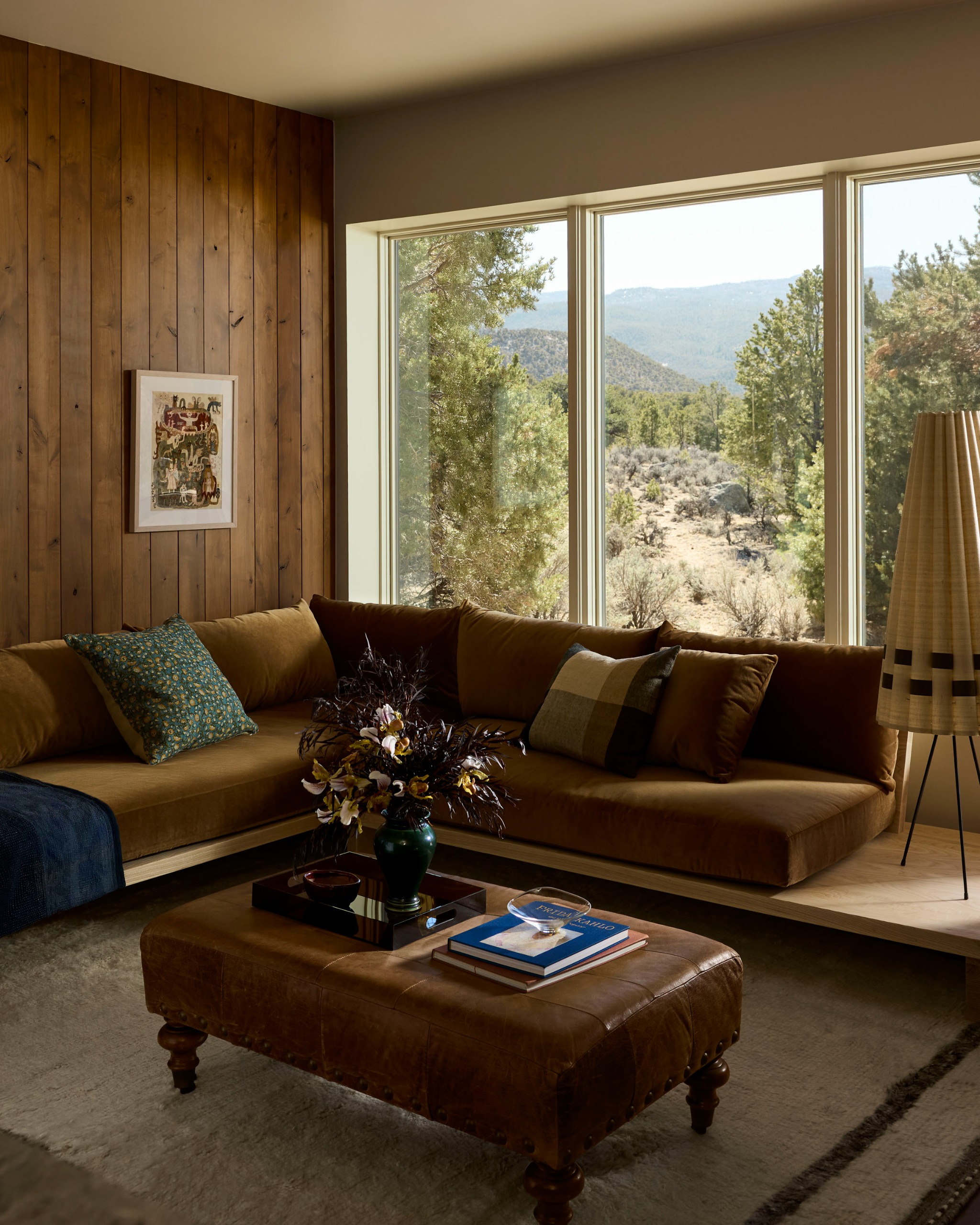 A sitting room with a built-in window seat with brown-ochre cushion seating in the corner by windows with sweeping views of the landscape with a leather pouffe in the center with decor on top