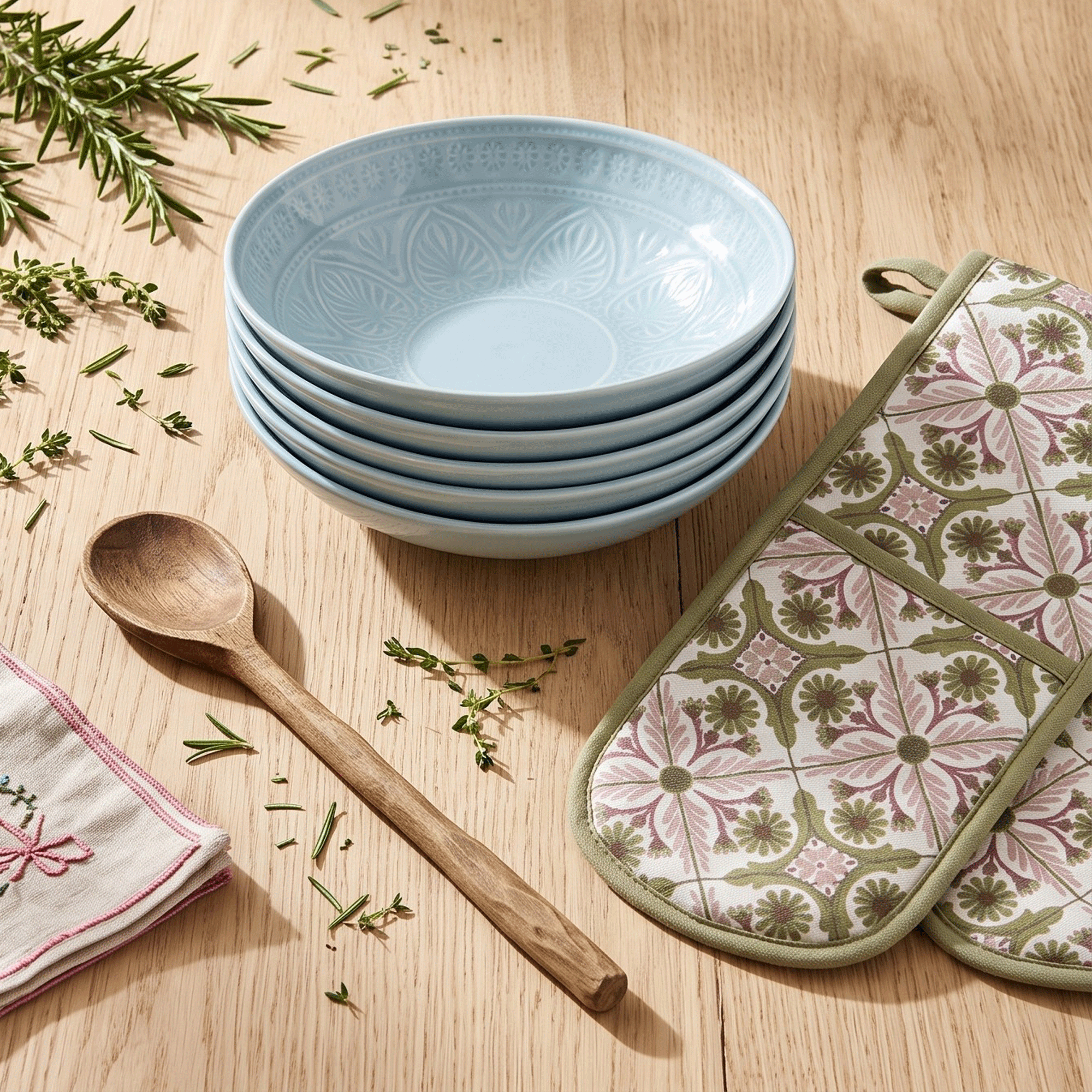 a wooden surface with a stack of blue bowls, a wooden spoon and a patterned oven mitt with scattered herbs