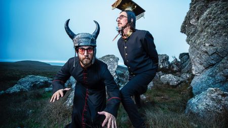 two men in black military tunics and horned hats cavort about in a rocky pasture