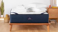 The best queen mattress of 2024 image shows The DreamCloud hybrid on a wooden bed frame in a white bedroom