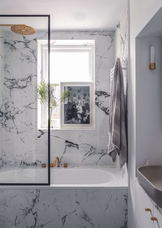 Bathroom with white and pale grey marbled walls and bath panel. Bath with gold coloured showerhead and window with a framed black and white photograph on the windowsill. A renovated penthouse duplex apartment in West London, home of Nick Prior and girlfriend Anna Lewis, designed by Amelie von Celsing.