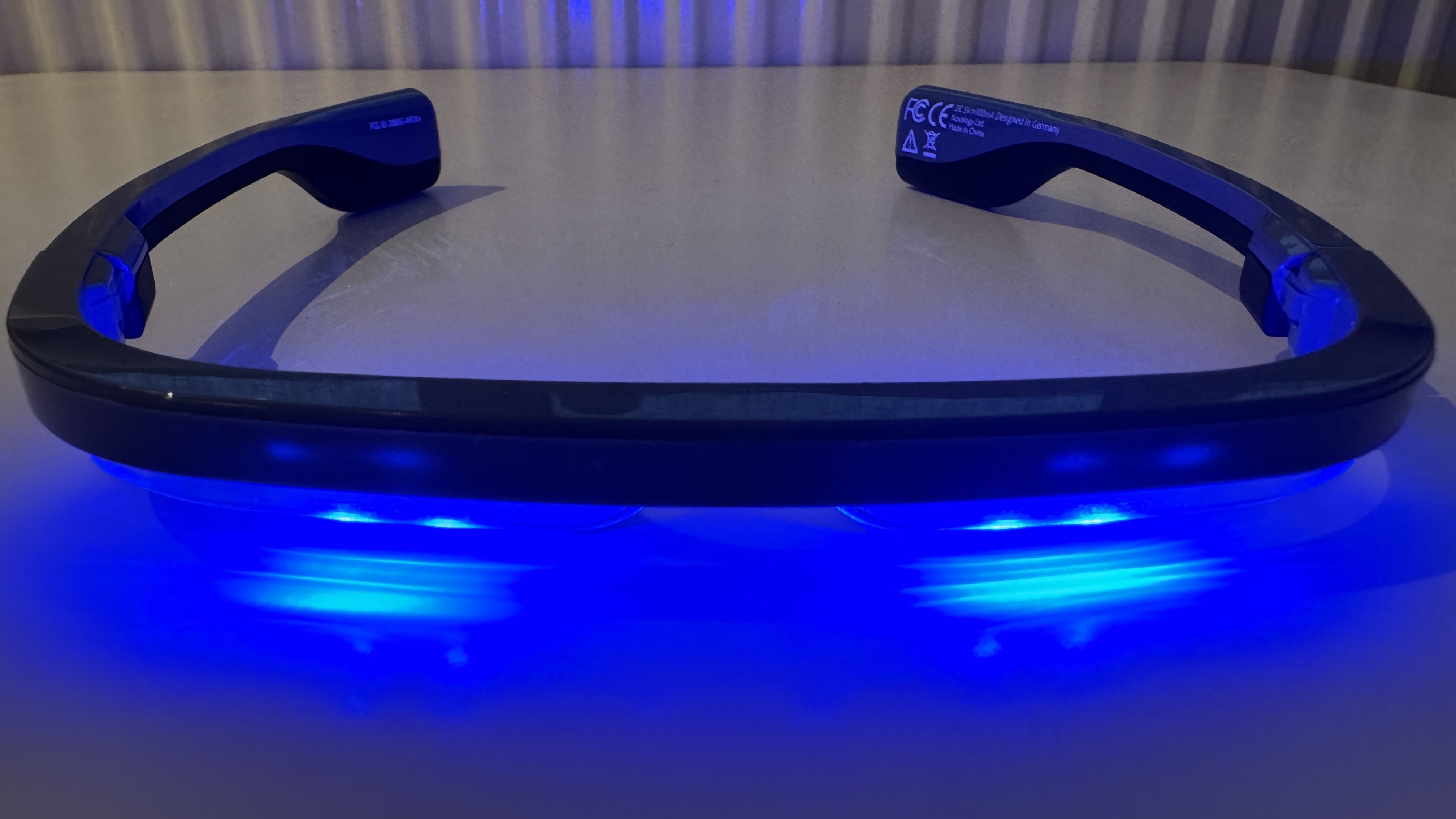 The AYO+ Blue Light Therapy Glasses switched on and placed on a white wooden surface so that the blue light diodes create a bright reflection
