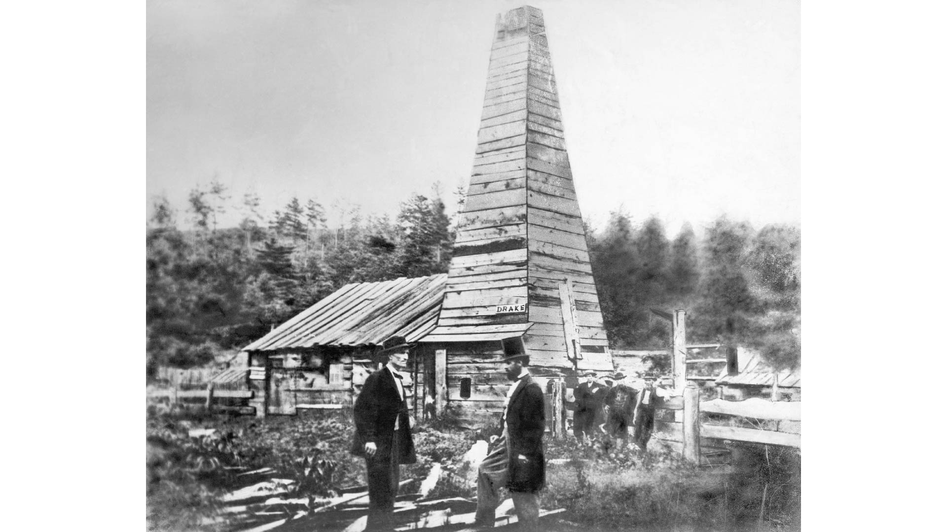 An old black and white photo of Edwin Drake's first oil well. The photo features men in suits and tophats standing in front of the well, and a large wooden structure