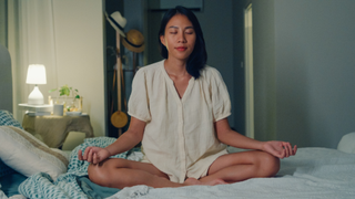 A woman sat cross legged in her pyjamas on her bed with her eyes closed and hands resting on each knee meditating