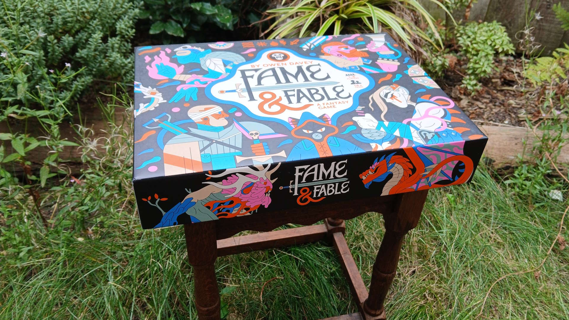 A boxed copy of Fame & Fable sat on a wooden bench outside amongst grass and plants