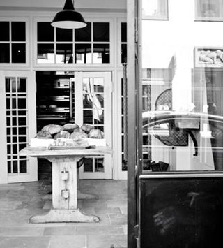 A black and white photo of a wooden table with loaves of bread stacked on top of it in front of a open wood and glass door.