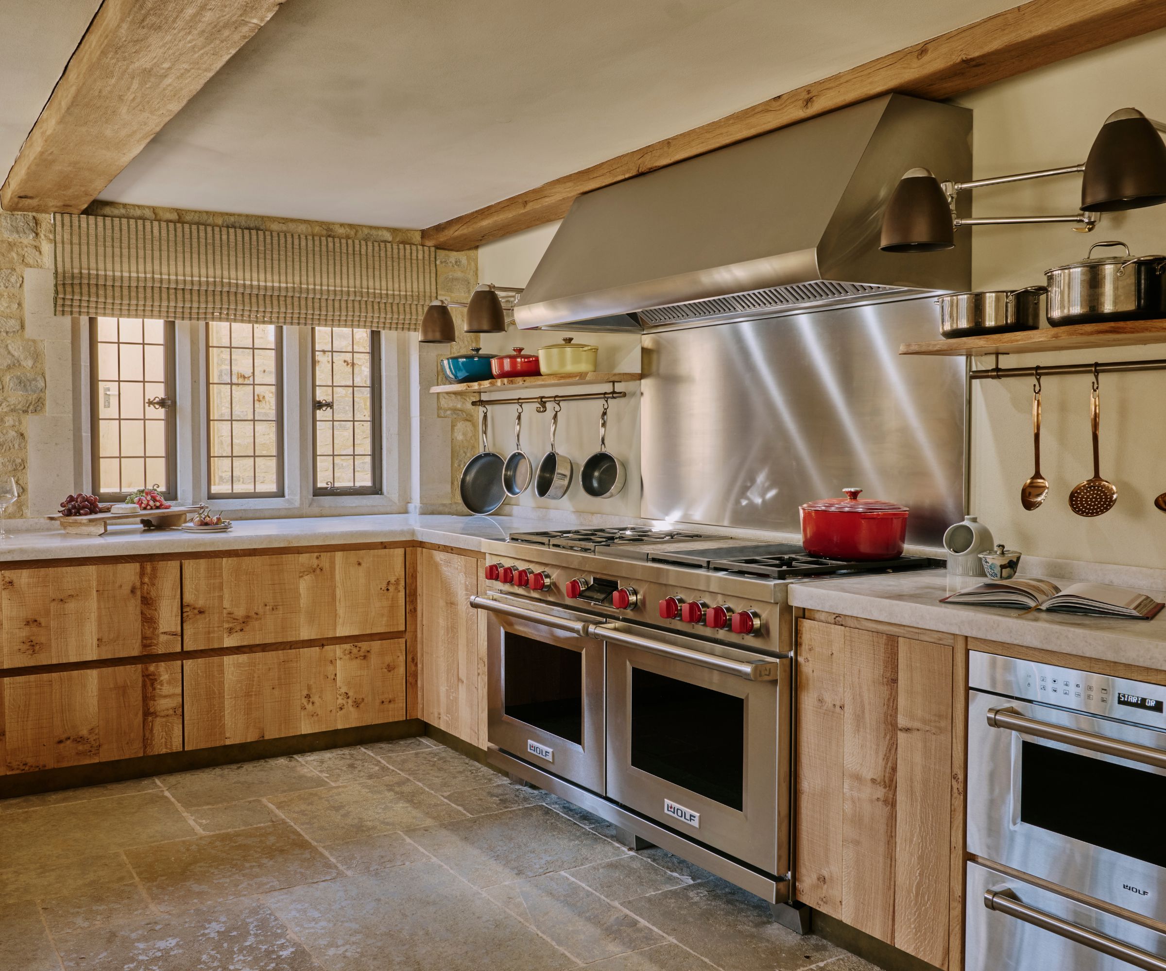 A Cotswolds kitchen with wooden cabinets, marble countertops, exposed stone walls, and stainless steel appliances and range hood