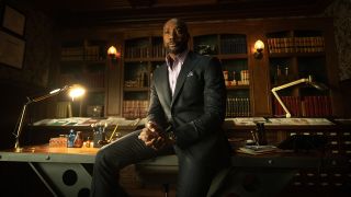 Watson sitting on his desk with pens and other desk items scattered about. A desk lamp is on. 