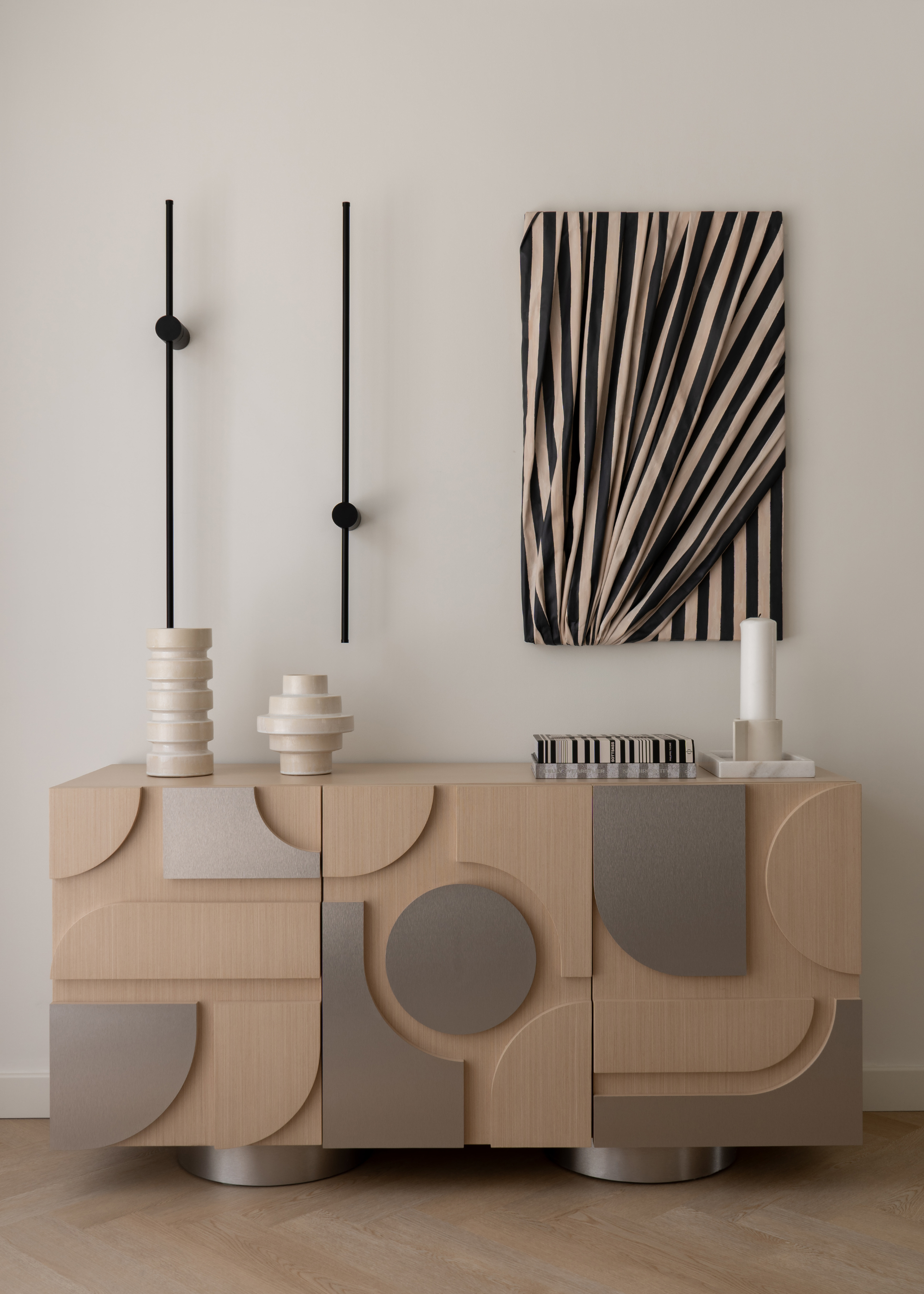 A living room with a chrome and wood sideboard holding stacked vases, books, and a large candle on a beige holder by black wall sconces and a draped art piece