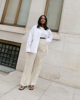 Die Mode-Influencerin Karina von Style Idealist posiert auf einem Bürgersteig in der Stadt und trägt eine Outfit-Formel aus einem weißen Basic-Top, einem weißen Button-Down-Hemd, einem schwarzen Gürtel, einer braunen Hose und schwarzen Flip-Flops mit Kitten-Heels