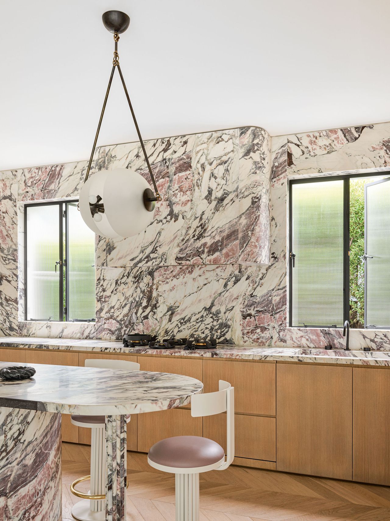 A marble covered kitchen with light toned wooden cupboards, two windows sit either side of the hob, and a large, modern chandelier above the kitchen island.
