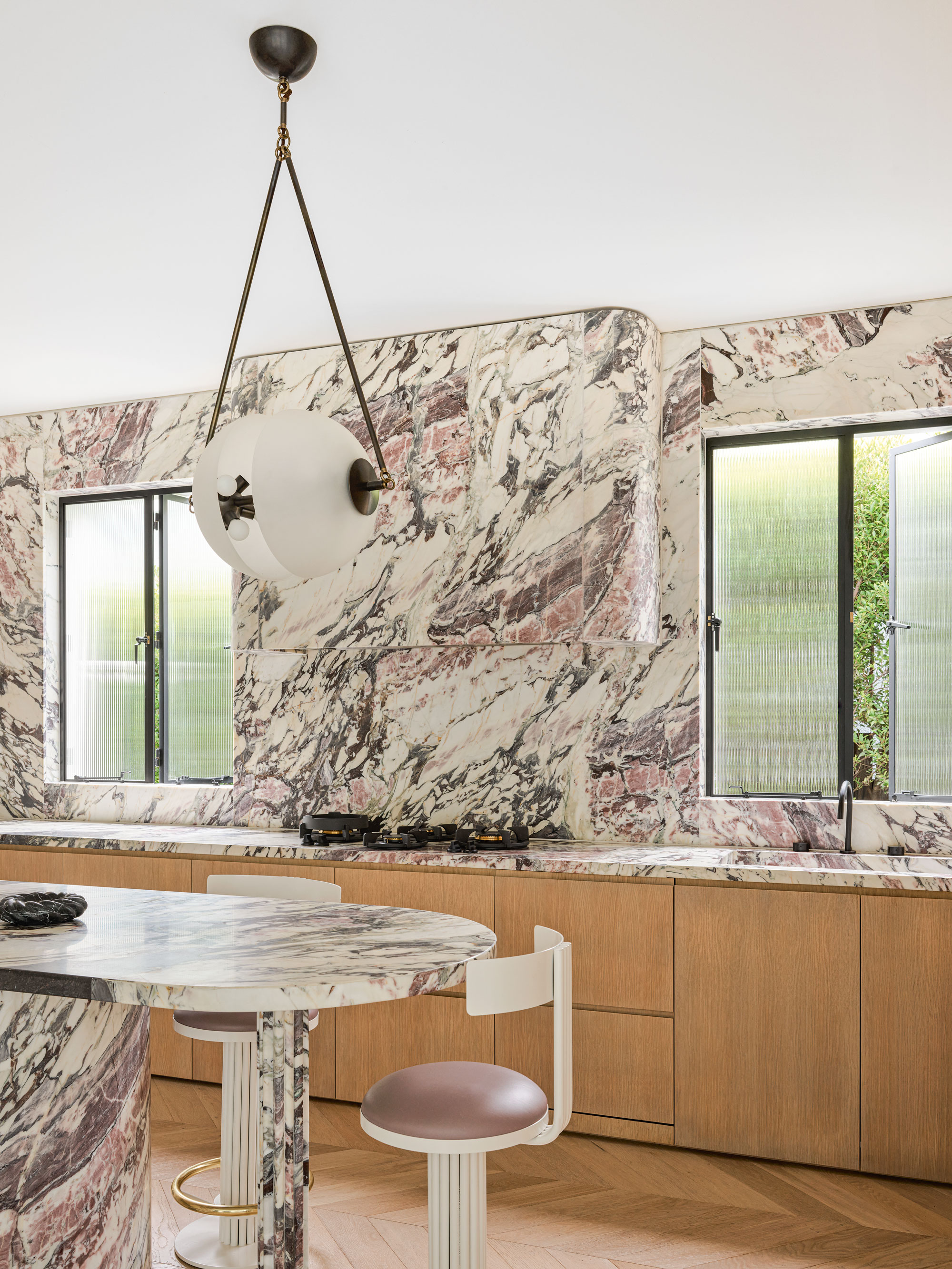 A marble covered kitchen with light toned wooden cupboards, two windows sit either side of the hob, and a large, modern chandelier above the kitchen island.