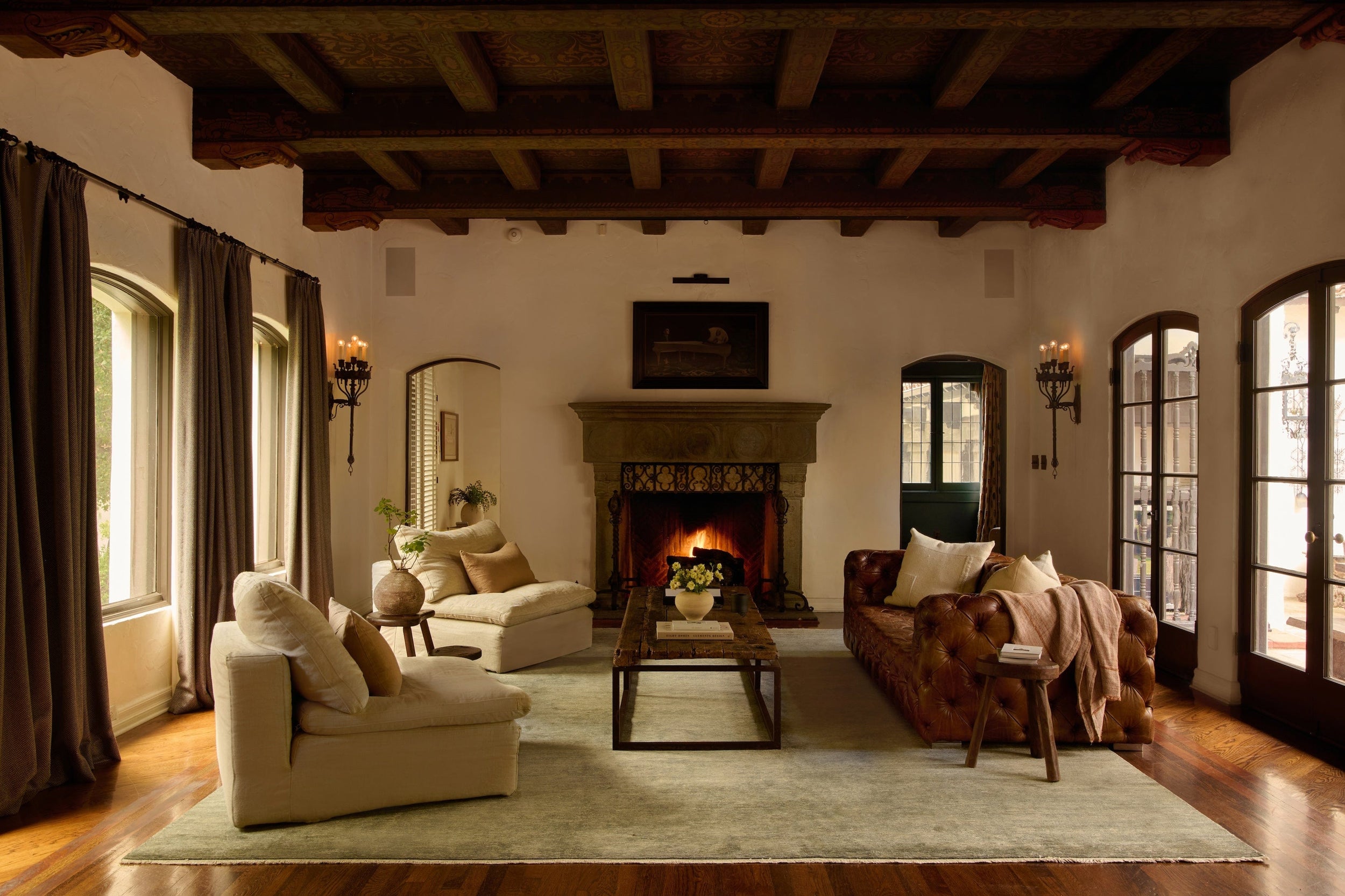 white living room with dark wood ceiling, grey rug on wooden floor, two white arm chair opposite a brown leather sofa. in between there is a wooden coffee table and behind there is a stone fireplace