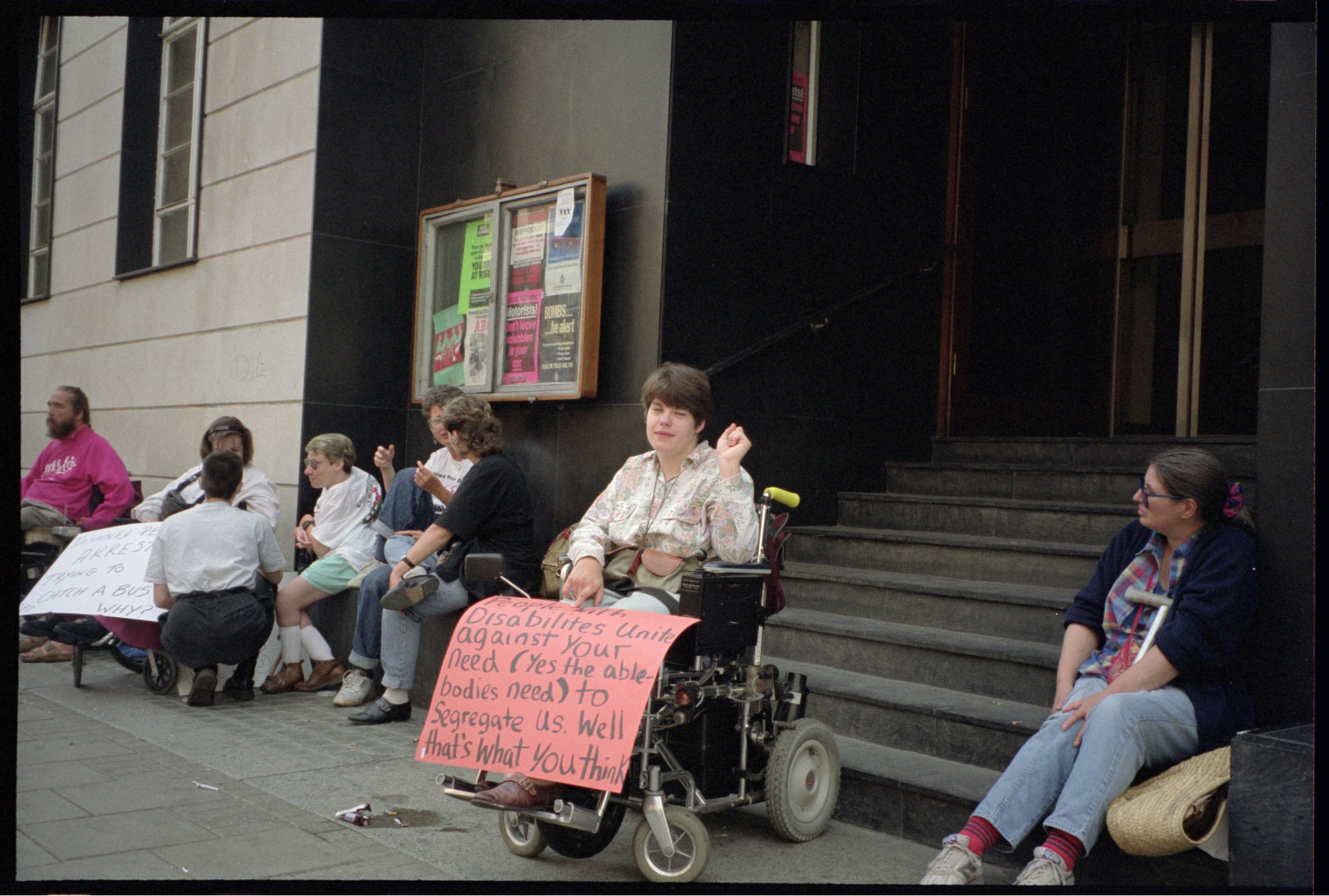 A smiling man in a wheelchair looks up defiantly while surrounded by Metropolitan Police officers, with a red London bus destination board for Trafalgar Square visible behind him.