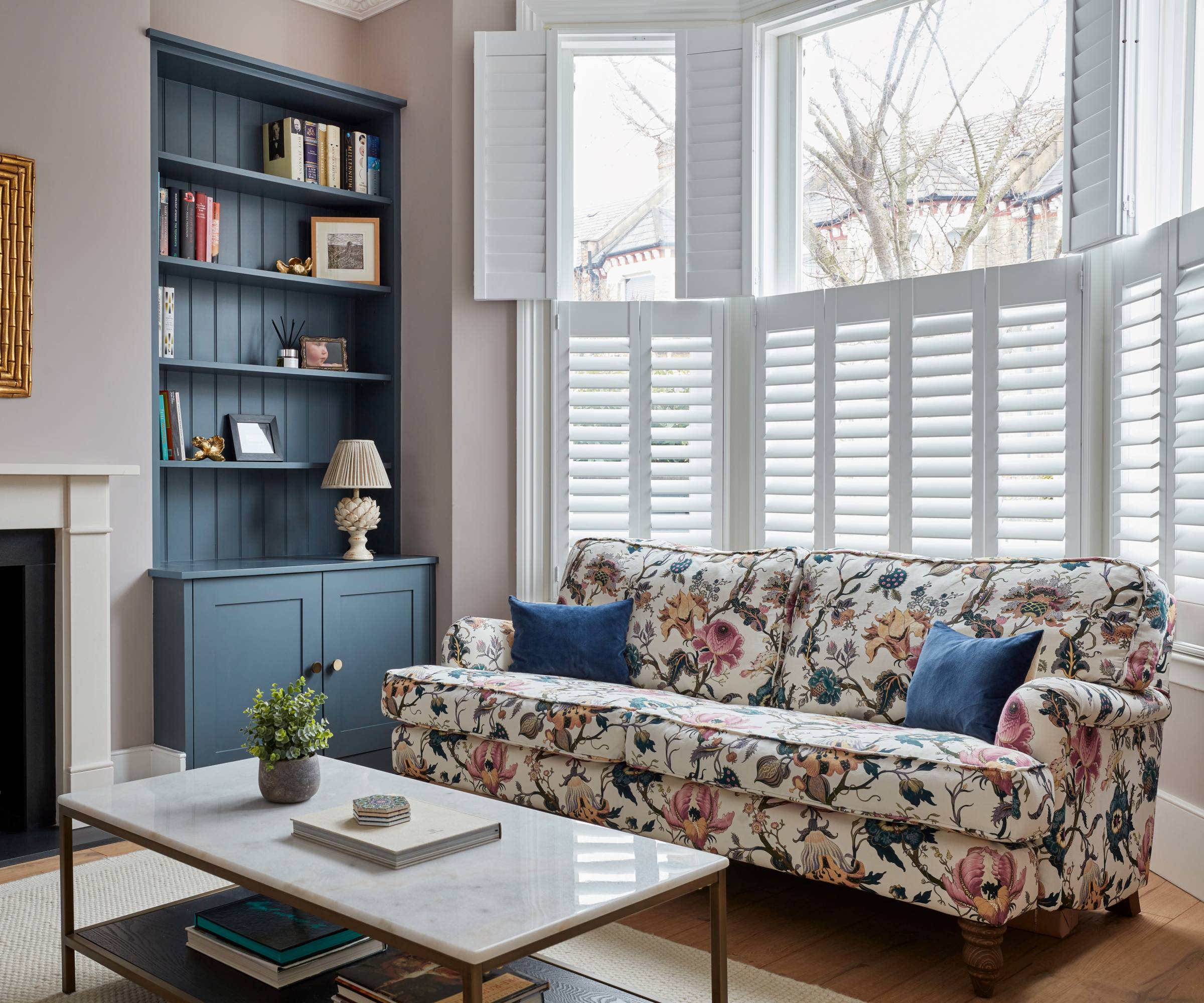 living room with split shutters in a bay window with the lower set closed and slats tilted, floral patterned sofa and blue built in alcove storage