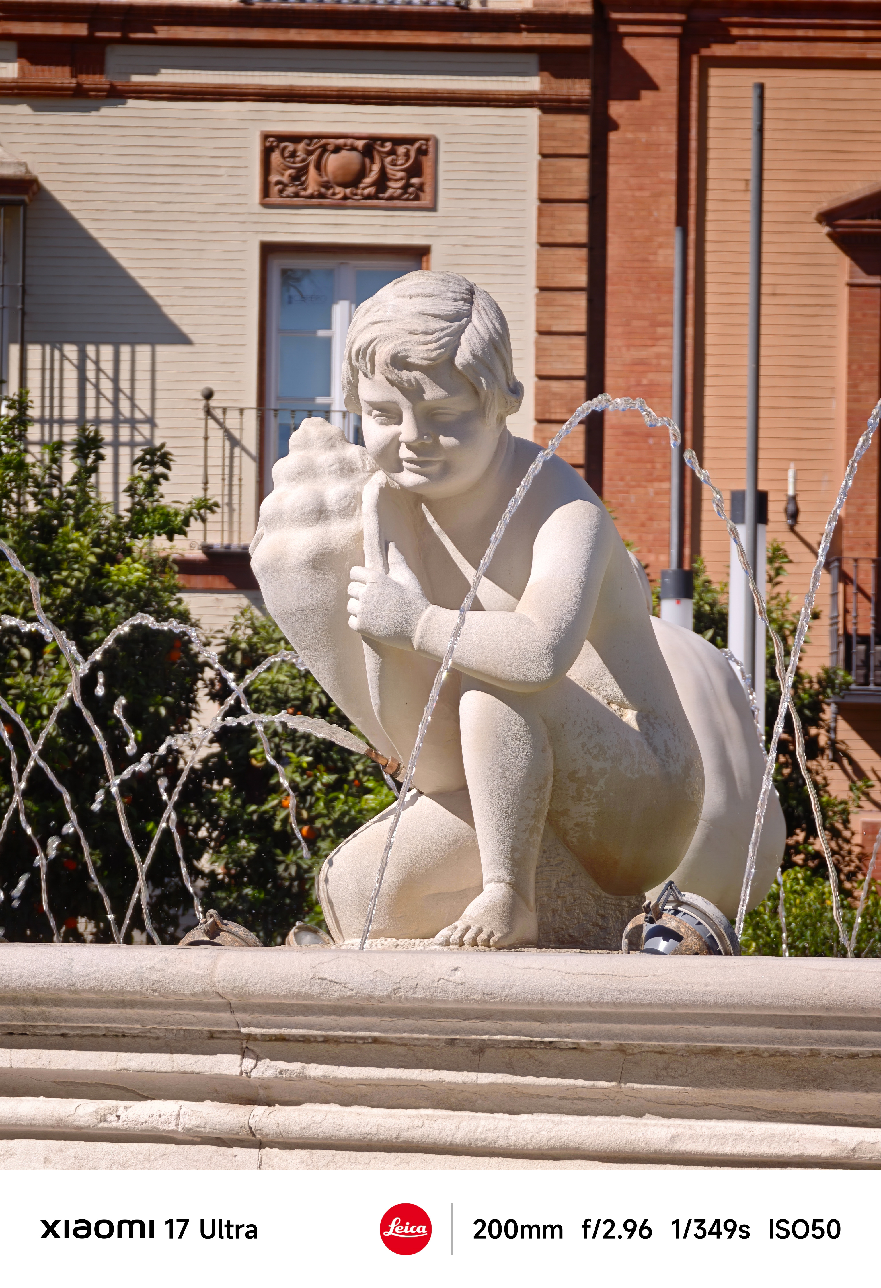 Stone statue of a child holding a shell in a fountain, water jets arcing around it with a brick building behind.