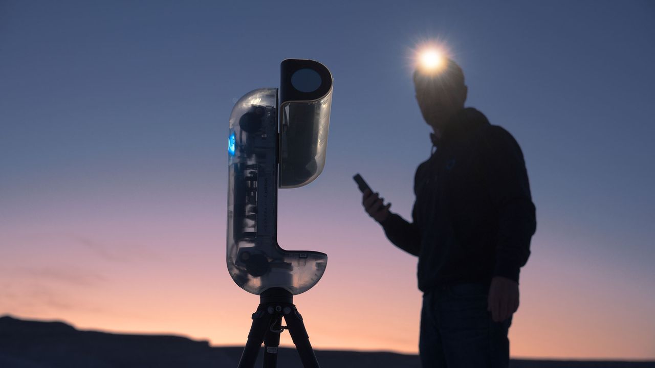 A man with a lit headtorch and smartphone stood next to the Vaonis Vespera II - X edition pointing at the sky with a sunset in the background.