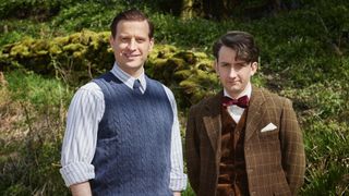 Nicholas Ralph in a blue sleeveless jumper and shirt as James stands next to James Anthony-Rose in a tweed jacket and bow tie as Carmody in All Creatures Great and Small.