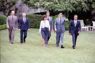 five men in suits and a woman wearing a skirt walk across a lawn