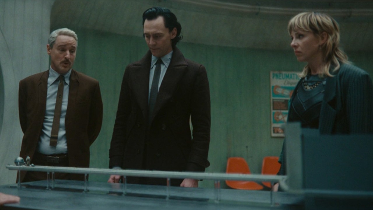 Image from the Marvel T.V. show Loki, season 2 episode 4. Three people are standing in front of a metal desk. On the left are two men wearing suits (the man on the left has short white hair and a moustache) (the man on the right is taller and has dark hair slicked back). To the right is a woman wearing a black dress and dark gray blazer with shoulder-length blonde hair). The three of them are staring down at the model on the table with concern.