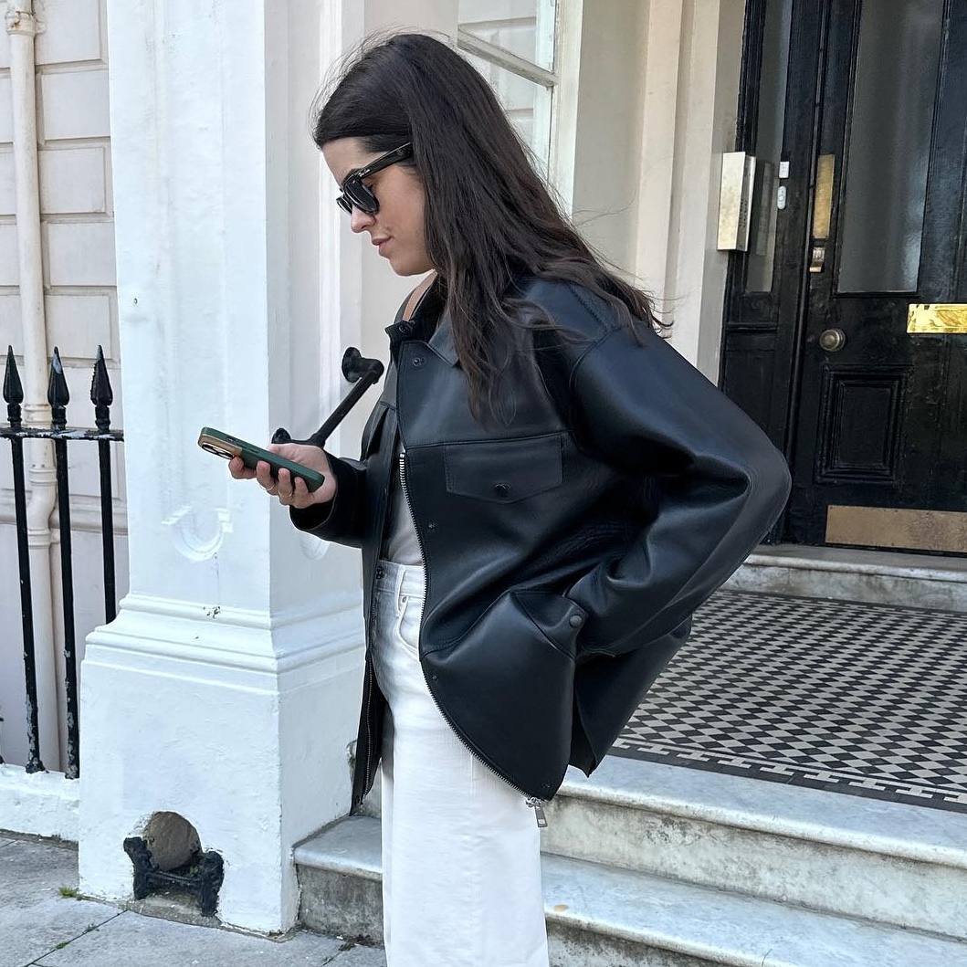 British style influencer Anna Howard poses on a London street looking at her phone wearing a black leather jacket, gray T-shirt, and white jeans