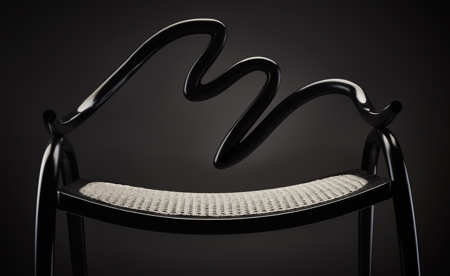 Front view of a black chair by Studio Swine featuring a curved back that resembles the shape of a snake and a woven seat in a contrasting colour. The chair is pictured against a dark background
