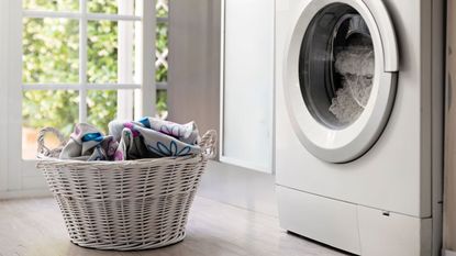 A white wicker laundry basket filled with bedding in front of a white, front-loading washing machine. 