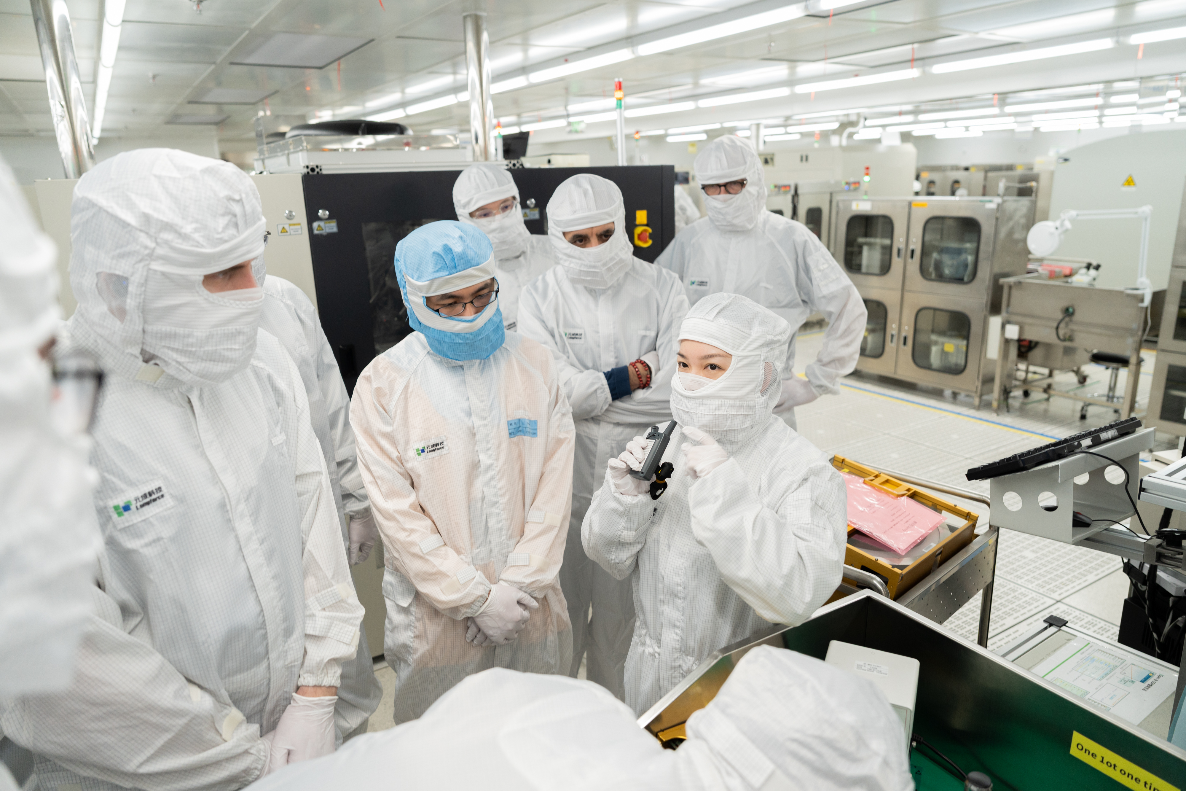 A group of people in cleanroom attire, including one wearing a blue headcover, are gathered for a discussion around a workstation in a bright, modern laboratory setting.