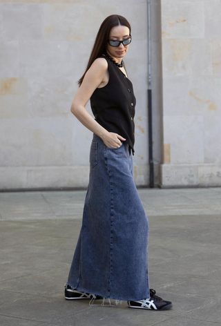 A woman wearing a long denim skirt with sneakers and black waistcoat.